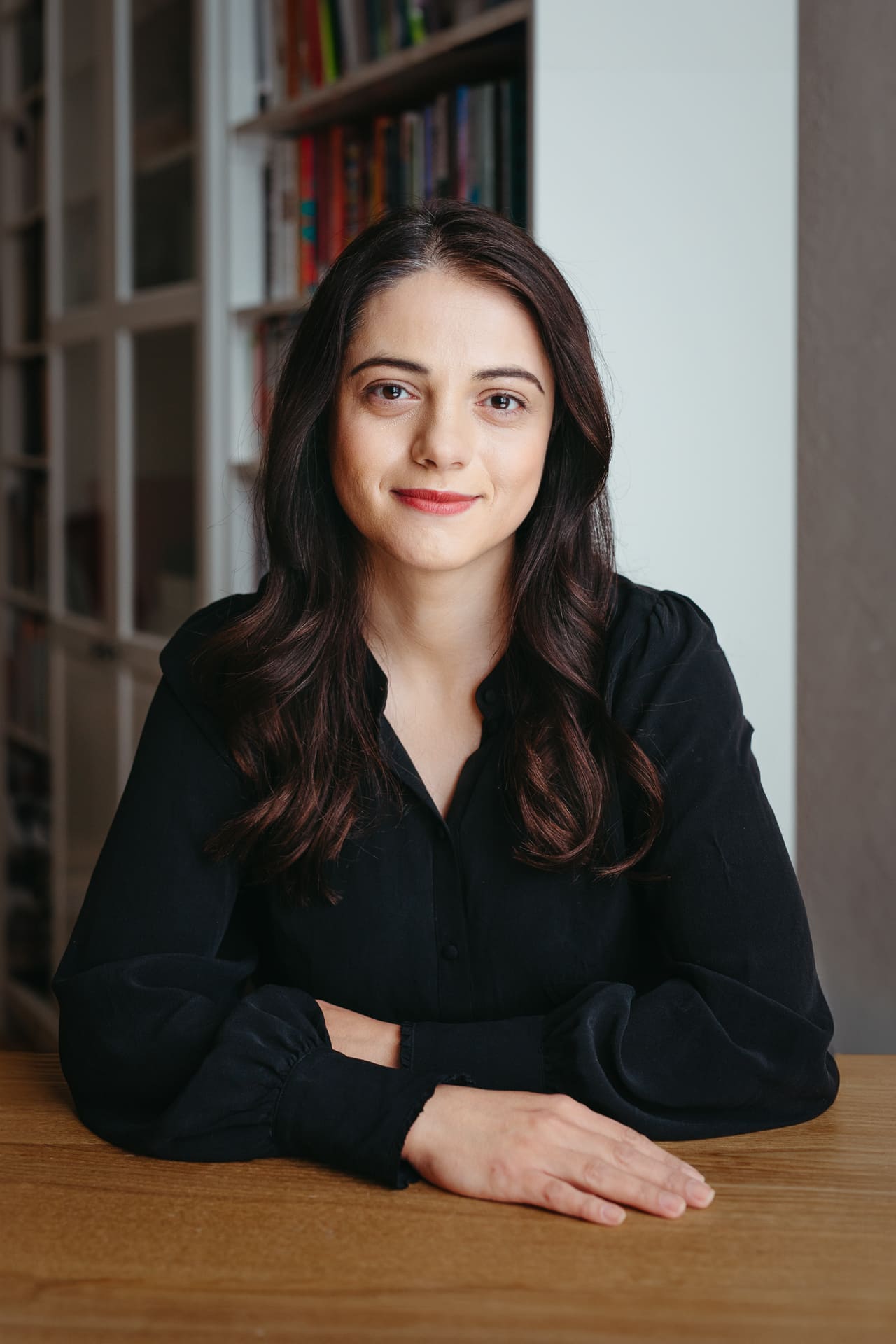 Portrait einer Frau am Tisch sitzend. Im Hintergrund ist ein Bücherregal zu sehen